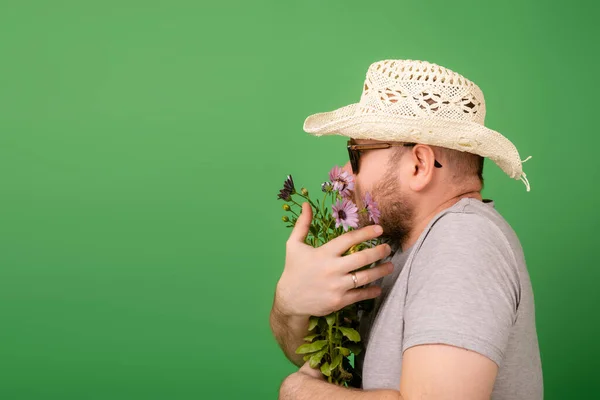 A male gardener, florist in a hat and sunglasses holds fresh flowers in pots on a green background. Plant breeding, sale.