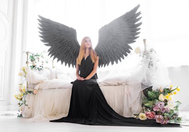 Beyaz bir yatakta, çiçeklerle kaplı siyah kanatlı bir melek. Genç düşünceli bir kız yatağın kenarında oturuyor..