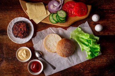 Hamburger ya da çizburger pişirmek. Klasik bir hamburger için farklı malzemeler. Izgara et, sebze, sebze, susamlı ekmeğin yanında soslar. Ahşap arkaplan.
