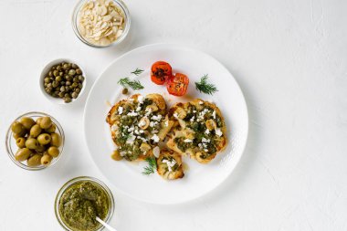 Baharatlı karnabahar bifteği, chimichurri sosu, badem gevreği, zeytin, kızarmış vişneli domates ve beyaz tabakta kapari. Vejetaryen yemeği. Beyaz arkaplan.