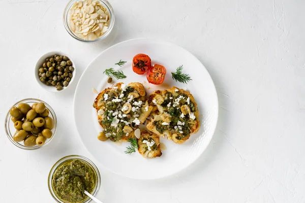 Baharatlı karnabahar bifteği, chimichurri sosu, badem gevreği, zeytin, kızarmış vişneli domates ve beyaz tabakta kapari. Vejetaryen yemeği. Beyaz arkaplan.