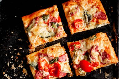 Dilimlenmiş kare pizza, turta, domatesli, sosisli, fırında pişmiş peynirli siyah bir fırın tepsisi..