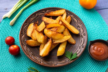 Tabakta büyük parçalar halinde patates kızartması. Masada turkuaz peçete ve farklı sebzeler var..