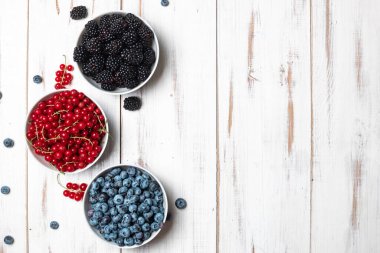 Taze böğürtlen, yaban mersini, böğürtlen ve beyaz ahşap arka planda bir tabakta kırmızı frenk üzümü. Metnin için yer.