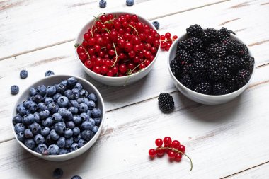Üç tabakta taze böğürtlen, böğürtlen ve beyaz ahşap arka planda kırmızı frenk üzümü. Meyve, dut arkaplan.