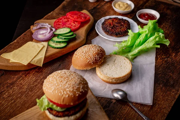 Cooking hamburger or cheeseburger. Different ingredients for a classic hamburger. Grilled meat, vegetables, greens, sauces near a sesame bun. Wooden background. Hamburger day.