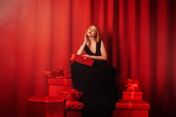 A joyful woman is sitting on a set of gifts. Middle-aged woman in a black dress. Red background. Happy face. Giving, receiving gifts concept.