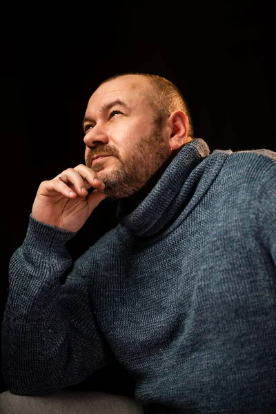 A middle-aged man in a sweater looks up. Unshaven man of normal appearance. Dark background. Thoughtfulness, meditation concept. Middle age crisis.