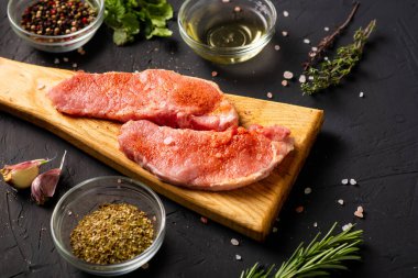 Kesme tahtasında çiğ et. Karanlık arka plan. Domuz eti pişirme hazırlığı. Yakınlarda çeşitli baharatlar ve baharatlar var..
