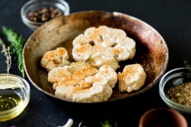 Karnabahar bifteği pişirme. Kızartma tavasına baharat serpiştirilmiş çiğ karnabahar. Zeytinyağı, bitkiler, yakınlarda çeşitli baharatlar. Koyu arkaplan.