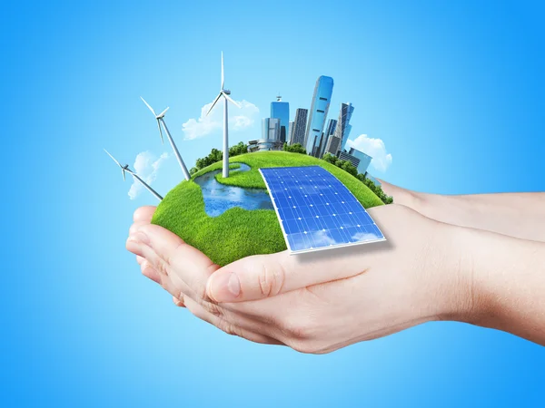 Manos sosteniendo prado verde claro con bloque de baterías solares, turbinas de molinos de viento y rascacielos de la ciudad — Foto de Stock