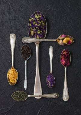 Siyah ve yeşil çay ile çeşitli kaşıklar, gül tomurcukları ve siyah arka planda mavi mallow çiçekleri ve taç yaprakları. Üst görünüm