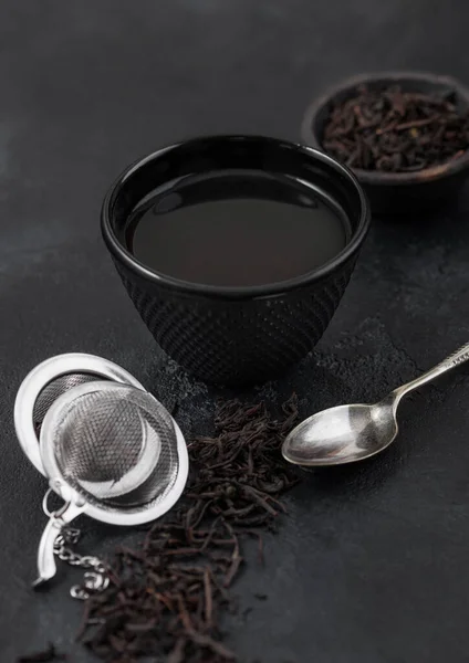Iron cup with black tea and strainer infuser with loose tea leaf on black with silver spoon. Top view