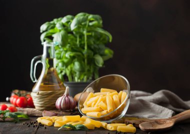 Cam kasede çiğ makarna, yağ ve sarımsak, fesleğen, biberli domates ve ahşap masa üzerinde keten havlu. Metin için boşluk