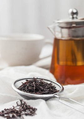 Strainer infusor with loose black tea on light cloth with clear glass teapot and white ceramic cup
