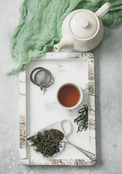 Green loose tea with tea ball strainer infuser and ceramic teapot with cup in wooden box with green cloth on light background