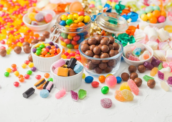Milk chocolate candies woth shell in jar with various jelly gums candies on white table with liquorice allsorts and strawberry bonbons with different sour sugar gums.