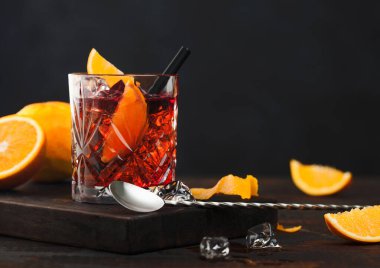Kristal bardakta Negroni kokteyli, portakal dilimi ve tahta üzerinde taze çiğ portakallar, ahşap arka planda kaşık..