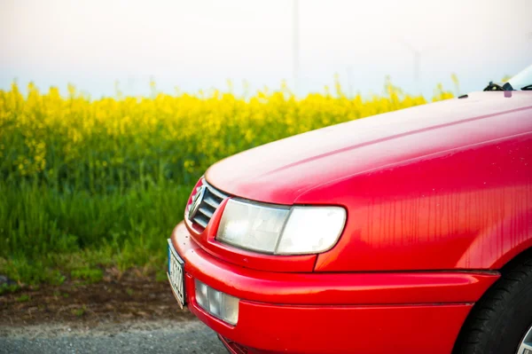 Das rote Auto steht neben dem Feld, auf dem der Raps blüht. und in der Ferne sieht man die Windräder — Stockfoto