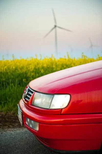 Červené auto stojí vedle pole kde znásilnit květiny. a v dálce vidíte větrné turbíny — Stock fotografie