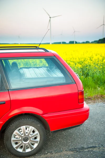 Red car stands next to the field where rape flowers. and in the distance you can see the wind turbines — Stock Photo, Image