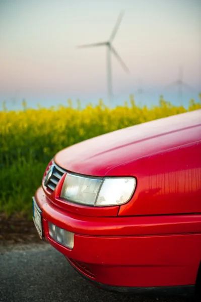 Červené auto stojí vedle pole kde znásilnit květiny. a v dálce vidíte větrné turbíny — Stock fotografie