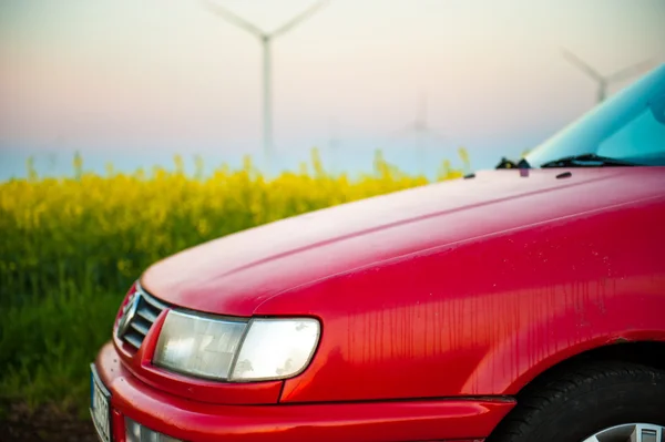 Červené auto stojí vedle pole kde znásilnit květiny. a v dálce vidíte větrné turbíny — Stock fotografie