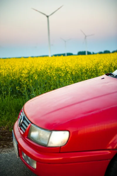 Красная машина стоит рядом с полем, где цветы рапса. и вдалеке вы можете увидеть ветряные турбины — стоковое фото