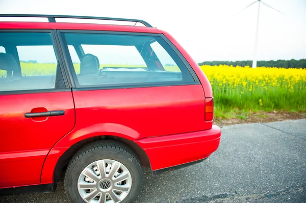 Carro vermelho fica ao lado do campo onde flores estupro. e na distância você pode ver as turbinas eólicas — Fotografia de Stock