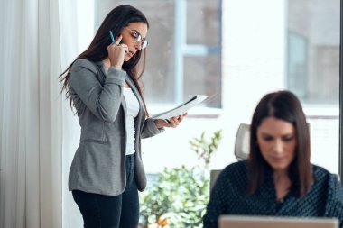 Ofisin penceresinin yanında cep telefonunu kullanan güzel bir kadının fotoğrafı..