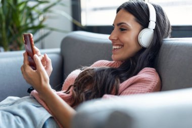 Evde kanepede otururken akıllı telefon kullanarak müzik dinleyen güzel bir kadın..