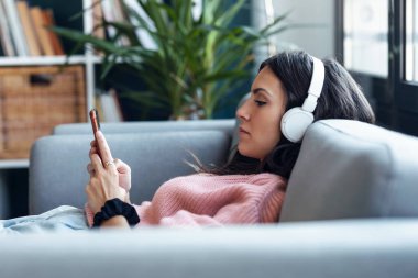 Evde kanepede otururken akıllı telefon kullanarak müzik dinleyen güzel bir kadın..