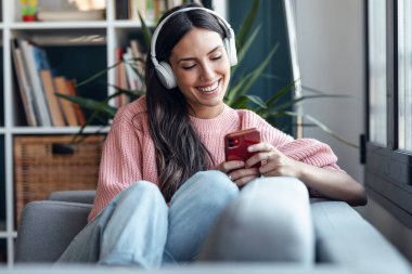 Evde kanepede otururken akıllı telefon kullanarak müzik dinleyen güzel bir kadın..