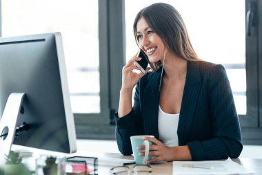 Ofiste bilgisayarla çalışırken çekici bir iş kadınının cep telefonuyla konuşması..