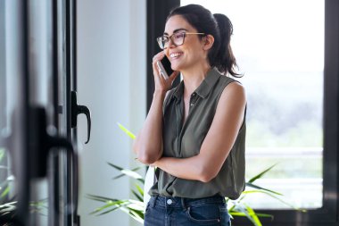 Ofisin penceresinin yanında cep telefonunu kullanan güzel bir kadının fotoğrafı..