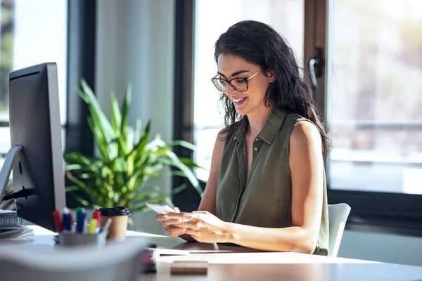 オフィスでコンピュータと仕事をしながら携帯電話を使って笑顔のビジネス女性のショット — ストック写真