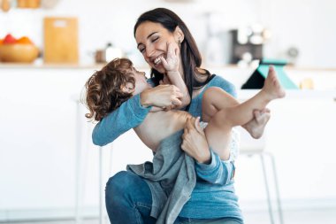 Güzel, genç bir annenin evdeki mutfakta oynaşıp, sevimli oğluna sarılmasının görüntüsü..
