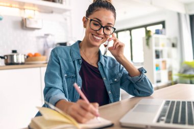 Evdeki mutfakta akıllı telefonla konuşurken dizüstü bilgisayarla çalışan gülümseyen bir iş kadını..
