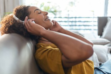 Evde kanepede oturup dinlenirken akıllı telefonuyla müzik dinleyen güzel, olgun bir kadının fotoğrafı..