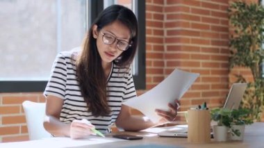 Evdeki ofiste bazı belgelere danışırken dizüstü bilgisayarla çalışan konsantre genç iş kadınının videosu..