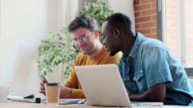 Modern başlangıç ofisinde el sıkışan bir şeyi kutlarken dizüstü bilgisayarla çalışan iki grafik tasarımcının videosu.