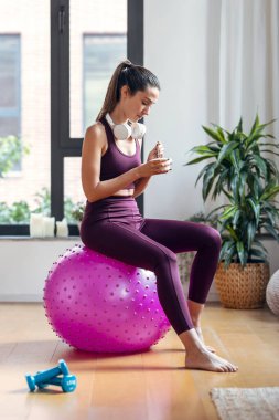 Evde fitness topunun üzerinde otururken yoğurt yiyen genç bir kadın..