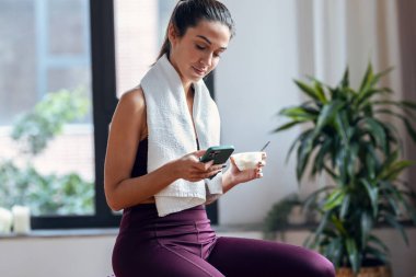 Spor yapan genç bir kadının yoğurt yerken akıllı telefonu evde fitness topunun üzerinde otururken görüntüsü..
