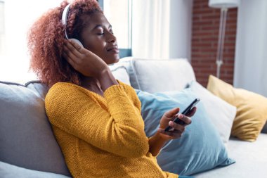 Evde kanepede otururken akıllı telefon kullanarak müzik dinleyen güzel bir kadın..