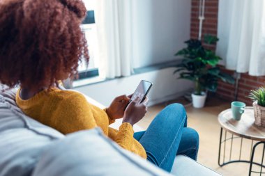 Evdeki kanepede otururken kendine güvenen genç bir kadının cep telefonunu kullanması..