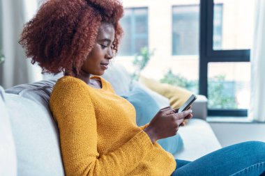 Evdeki kanepede otururken kendine güvenen genç bir kadının cep telefonunu kullanması..