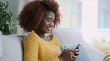 Gülümseyen genç bir kadının evdeki koltukta otururken cep telefonunu kullanmasının videosu..