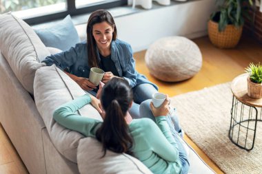 Oturma odasındaki kanepede oturmuş kahve içerken gülümseyen iki genç kadının görüntüsü..