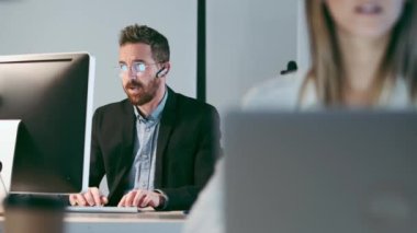 Yetişkin iş adamının bluetooth giydiği video modern başlangıç ofisinde meslektaşıyla bilgisayar üzerinde çalışırken çekilmiş..