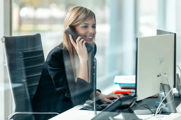 現代のスタートアップオフィスでオフィスの電話と話をしながら コンピュータを操作する美しい若いビジネス女性のショット — ストック写真